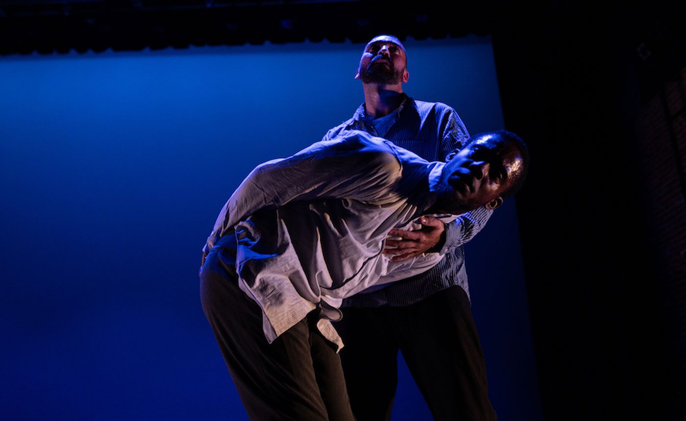 Two male dancers perform a dramatic duet on a dimly lit stage, with one leaning back dramatically into the other's embrace.