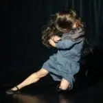 A woman in a blue dress performs a dynamic dance move on stage, her hair flung wildly covering her face.