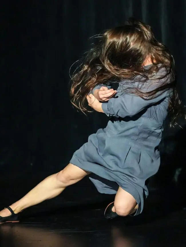 A woman in a blue dress performs a dynamic dance move on stage, her hair flung wildly covering her face.