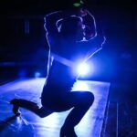 A silhouette of a dancer on stage, highlighted by a bright blue backlight, capturing a dynamic pose with one knee bent and hands above the head.