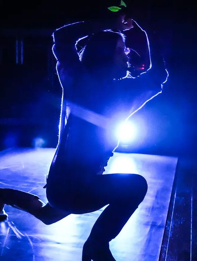 A silhouette of a dancer on stage, highlighted by a bright blue backlight, capturing a dynamic pose with one knee bent and hands above the head.