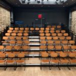 Small, empty theater with rows of orange chairs facing a black stage, flanked by exposed brick walls and dark curtains.