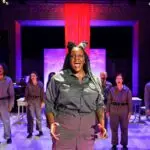 A woman stands center stage singing expressively with a cast in uniform behind her, all set against a theatrical backdrop with a red curtain.