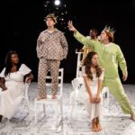 Five actors in eclectic costumes perform on a stage with a white floor, using chairs as props in a dramatic scene.