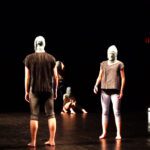 Contemporary dance performance featuring several dancers in casual attire with masked faces on a dimly lit stage.