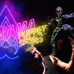 A man is holding a puppet in front of a neon sign.