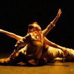 Two dancers in a dramatic pose on a dimly lit stage, one is arching back while the other stretches out an arm, both are wearing flowing costumes.