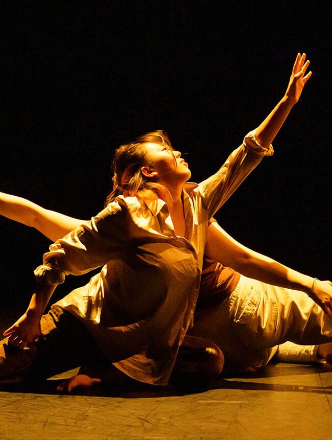 Two dancers in a dramatic pose on a dimly lit stage, one is arching back while the other stretches out an arm, both are wearing flowing costumes.