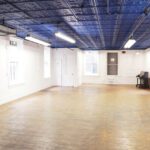 Empty dance studio with wooden floors, mirrored wall on the left, white walls, and a blue decorative ceiling. there is a piano in the corner.