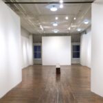 Empty art gallery with white walls, spotlights, a wooden bench in the center, and a large opaque panel at one end.